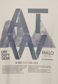 T-shirt w kolorze off-white z dużym, ciemnoniebieskim graficznym napisem: "ATW," "OFF DUTY GEAR" oraz informacjami na temat samolotu B-583 w nowoczesnej czcionce.
