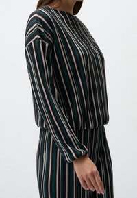 Femme portant un haut à manches longues et un pantalon avec des rayures verticales noires, blanches et vertes sur un fond clair uni.