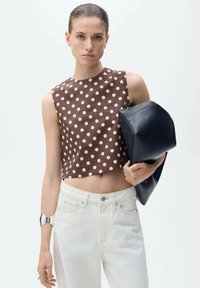 Hnědý crop top s puntíky je kombinován s bílými džíny s vysokým pasem. Modelka drží v jedné ruce černou, strukturovanou kabelku a na zápěstí má metalickou náramkovou. 