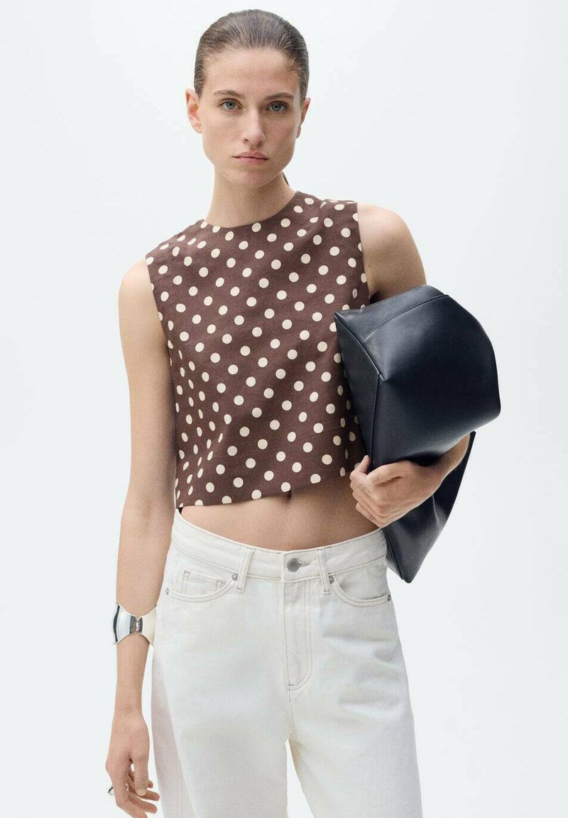 Hnědý crop top s puntíky je kombinován s bílými džíny s vysokým pasem. Modelka drží v jedné ruce černou, strukturovanou kabelku a na zápěstí má metalickou náramkovou. 