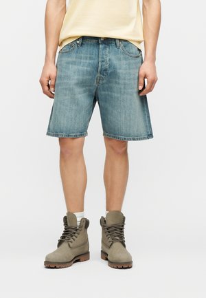 Άνδρας που φοράει ξεθωριασμένα μπλε τζιν σορτς, μπεζ μπλουζάκι, λευκές κάλτσες και μπότες αστραγάλου σε χρώμα ταφ με κορδόνια, στέκεται μπροστά σε έναν απλό φόντο.