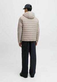 Beige Daunenjacke mit Kapuze, ausgestattet mit einem gesteppte Design und weicher Textur. Kombiniert mit dunklen, weit geschnittenen Hosen.