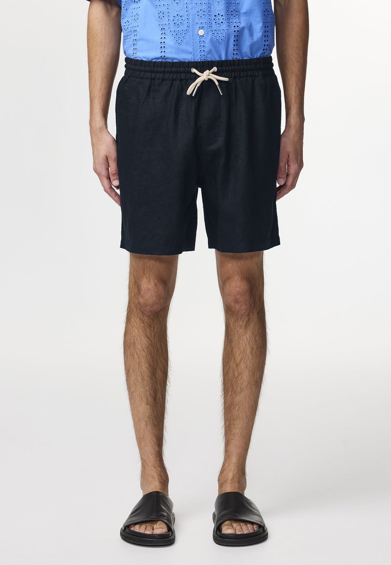 Pantaloni corti neri con cintura elastica e cordoncino beige. Indossati con una camicia button-down blu e sandali neri a infradito.