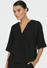 Blusa nera a V con maniche corte, dettagli arricciati sotto il colletto, realizzata in un tessuto morbido, vestibilità rilassata e trama discreta.