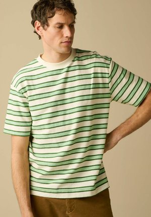 Homme portant une chemise blanche à manches courtes avec des rayures horizontales vertes, pantalon marron, regardant vers le bas avec la main sur la hanche sur un fond beige.