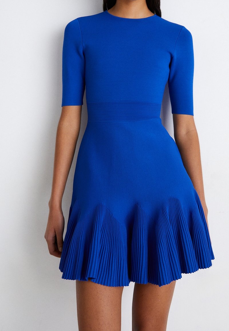 Femme portant une robe ajustée bleu vif à manches courtes, avec une taille côtelée et une jupe plissée, debout sur un fond blanc.