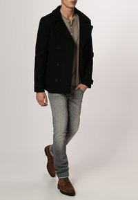 Cappotto peacoat in lana nera con bottoni doppi, abbinato a una maglietta henley grigia chiara, jeans grigi e scarpe in suede marroni.