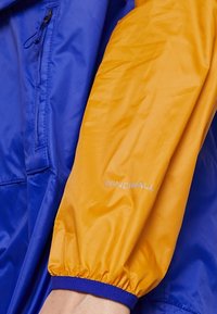Vindtät jacka i blå och orange nylon. Har elastiska ärmmuddar, en dragkedjeförsedd ficka och en "WINDWALL" logotyp på ärmen.