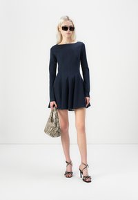Marineblauwe jurk met lange mouwen en een uitlopende rok, gemaakt van een gestructureerde stof, gecombineerd met een handtas van slangenleer en zwarte hakken met bandjes. Model draagt een zonnebril.