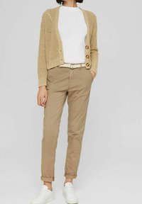 Vrouw draagt een beige gebreid vest, wit overhemd, beige broek met riem en witte sneakers, staand met handen in de zakken.