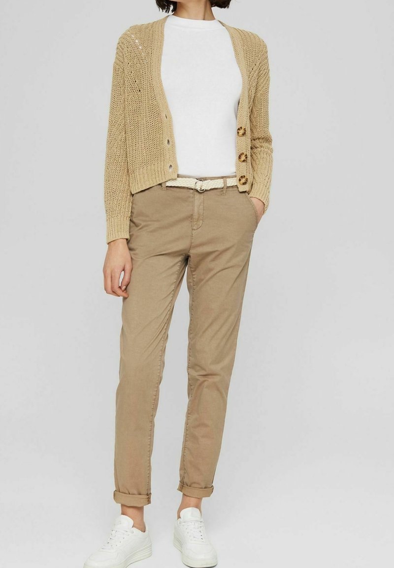 Vrouw draagt een beige gebreid vest, wit overhemd, beige broek met riem en witte sneakers, staand met handen in de zakken.
