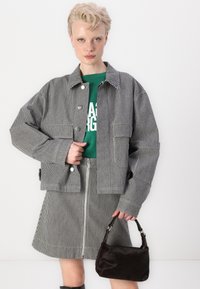 Chaqueta recortada a rayas en blanco y negro con una falda a juego. Camiseta gráfica verde debajo. Sosteniendo un pequeño bolso marrón oscuro.