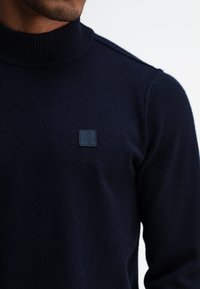 Marineblauer Strickpullover mit hohem geripptem Kragen und kleinem strukturiertem Logopatch auf der Brust. Der Stoff hat eine weiche, glatte Textur.