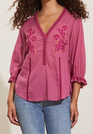 Vrouw draagt een roze geborduurde blouse met driekwart mouw met ruches en blauwe spijkerbroek, weergegeven van schouders tot heupen.