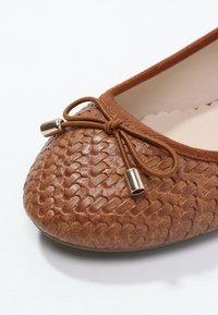 Ballerine marron tissée à texture avec un petit nœud et des embouts métalliques sur un fond clair uni.