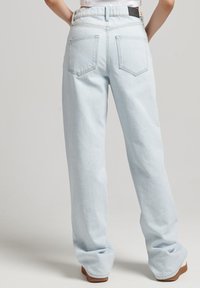 Persona vestida con jeans rectos de color azul claro y zapatos marrones, de pie con las manos en las caderas contra un fondo neutro.