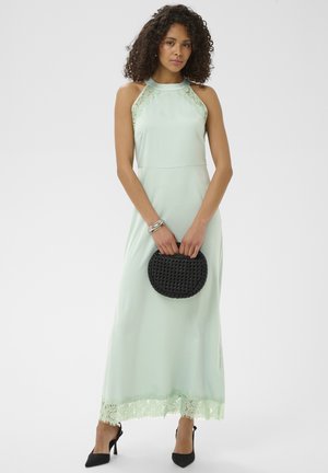 Kobieta o kręconych włosach, ubrana w jasnozieloną, bezrękawnikową sukienkę maxi z koronkowymi detalami, trzymająca czarną plecioną torebkę i nosząca czarne szpilki.