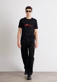 Svart t-skjorte med grafikk av et løpende dyr i rødt, parret med svarte cargo-bukser og svarte joggesko. Modellen har på seg mørke solbriller.