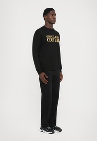 Črna bombažna majica s kapuco z zlatim logotipom "VERSACE JEANS COUTURE", v kombinaciji s črnimi hlačami in črnimi športnimi copati. Preprost dizajn, klasičen kroj.