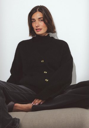 Čierny oversized kardigan so zlatými gombíkmi, nosený cez čierny rolák. Mäkká textúra, voľný strih a dlhé rukávy.
