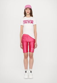 Bílé tričko s krátkým rukávem a růžovým logem "Versace Jeans Couture", doplněné o lesklé růžové cyklistické šortky a bílé tenisky. Součástí je také růžová kšiltovka.
