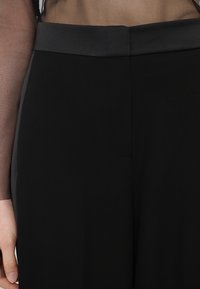 Pantalons noirs taille haute avec une texture lisse et un design épuré, présentant un bandeau en satin à la taille et des coutures subtiles.