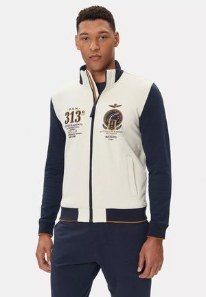 Giacca a track con zip in crema e blu marino, con logo ricamato, accenti dorati, colletto alto e tasche laterali. Tessuto morbido e design aderente.