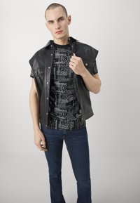 Chaleco de cuero negro sin mangas sobre una camiseta gráfica negra con texto blanco. Jeans azul oscuro. El modelo está de pie de frente con la mano en el chaleco.