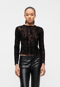 Γυναίκα με πλεκτά μαλλιά που φοράει διάφανο μαύρο τοπ με μοτίβο φίδι και μακρύ μανίκι και μαύρο δερμάτινο παντελόνι, στέκεται μπροστά σε λευκό φόντο.