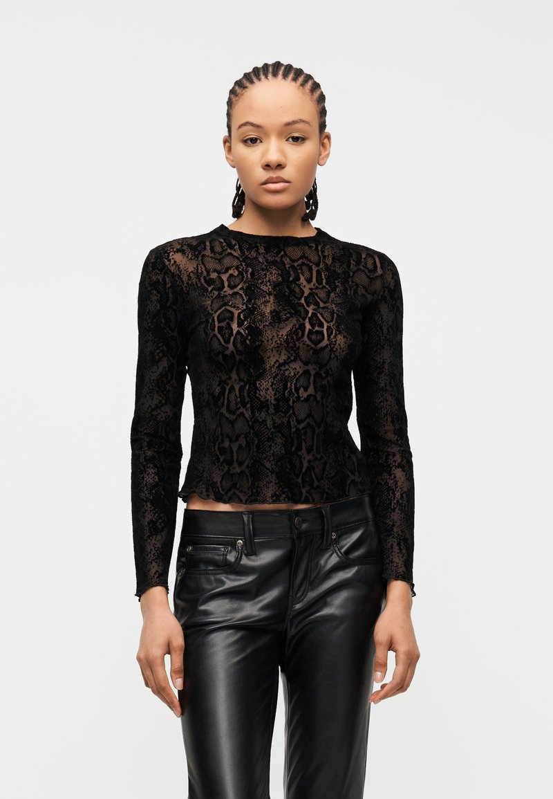 Γυναίκα με πλεκτά μαλλιά που φοράει διάφανο μαύρο τοπ με μοτίβο φίδι και μακρύ μανίκι και μαύρο δερμάτινο παντελόνι, στέκεται μπροστά σε λευκό φόντο.