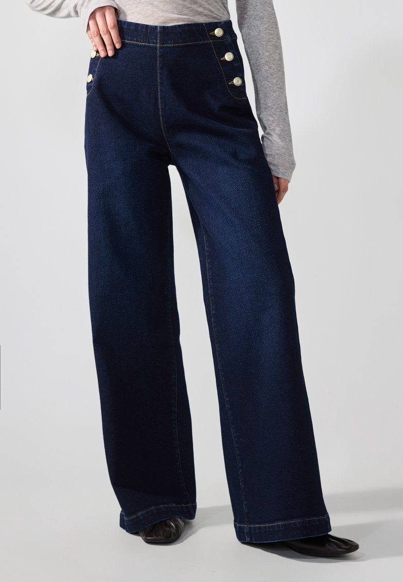 Jean denim bleu foncé à jambes larges avec trois boutons décoratifs sur chaque hanche, porté avec un haut gris à manches longues et des chaussures noires.