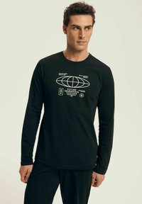 Man wearing black long-sleeve shirt with white futuristic text and globe graphic, standing against a light background, looking sideways.