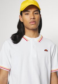 Biała koszulka polo z czerwonym i żółtym paskiem przy kołnierzy, z małym haftowanym logo na klatce piersiowej. Żółta czapka dopełnia strój.