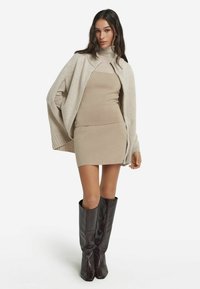 Beige vest van een wolmix met open voorkant, gedragen over een aansluitende beige jurk. Het model draagt donkerbruine leren laarzen tot over de knie.