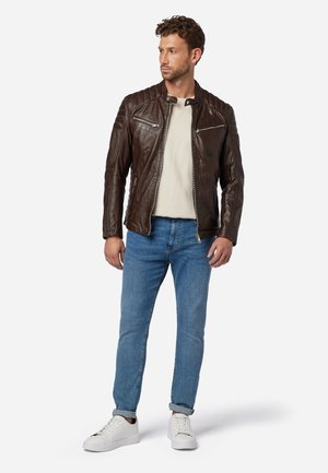 Brown leather jacket with quilted shoulder details, two zippered chest pockets, and a mandarin collar, worn over a cream shirt and blue jeans.