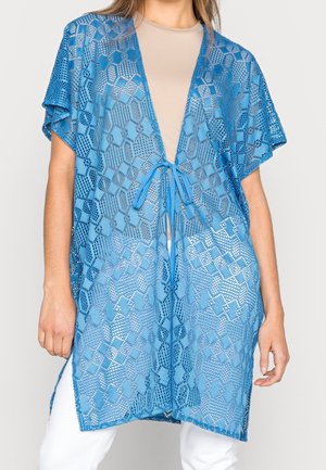 Cardigan bleu ouvert en crochet avec motifs géométriques, manches courtes et fermeture à nouer porté sur un haut beige et un pantalon blanc.