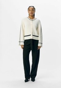 Cardigan a coste crema con accenti neri, bottoni perlati e maniche larghe, abbinato a pantaloni neri a vita alta e scarpe a punta.
