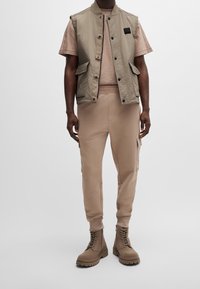 Ležérní outfit zahrnující světle béžovou bezrukávou vestu, béžovou košili s krátkým rukávem, související joggingové kalhoty a hnědé šněrovací boty.