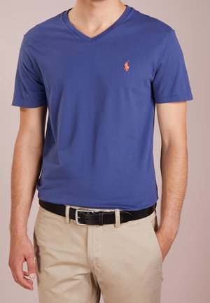Mężczyzna w niebieskim T-shircie z dekoltem w serek z pomarańczowym logo polo oraz beżowych spodniach z czarnym paskiem, z rękami swobodnie opuszczonymi wzdłuż ciała.