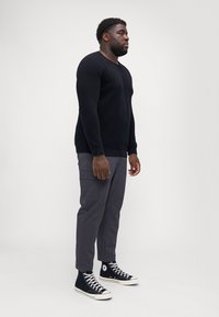 Pulover lung negru cu mâneci, pantaloni cargo gri cu buzunare laterale și pantofi sport negri cu glezna înaltă și accente albe, prezentate pe un fundal simplu.