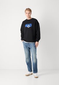 Crna majica s kapuljačom s plavim "HUGO" grafičkim natpisom i kružnim dizajnom sprijeda, uparena sa svijetloplavim trapericama i bijelim tenisicama.