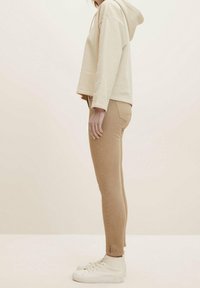 Sudadera corta beige con capucha, combinada con vaqueros ceñidos color marrón claro. El modelo lleva zapatillas altas blancas. Diseño simple con detalles de costura sutiles.