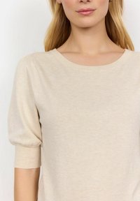 Pull en tricot beige avec un col rond et des manches bouffantes courtes. Le tissu semble doux et léger, idéal pour une tenue décontractée.