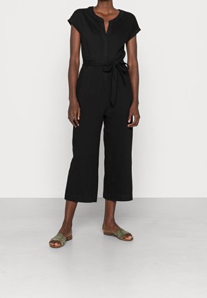 Femme portant une combinaison noire à manches courtes avec une ceinture à la taille et des jambes larges et courtes, associée à des sandales plates vert olive.