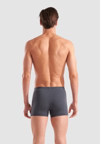 Grå boxershorts med en slät textur, som har en åtsittande passform och elastisk midja, sedd bakifrån mot en ljusgrå bakgrund.
