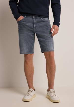 Mann trägt ausgewaschene graue Jeansshorts, navyblaues Langarmshirt und weiße Turnschuhe, steht mit einer Hand in der Tasche.