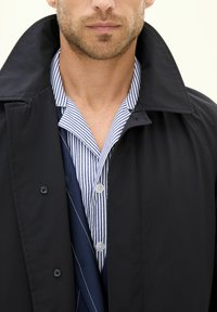 Schwarze Außenjacke mit hohem Kragen, über einem blau-weiß gestreiften Hemd und einer marineblauen Krawatte getragen, mit Knopfdetails und glatter Textur.