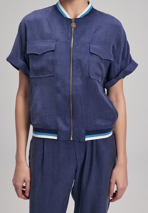 Femme portant une veste zippée bleu marine à manches courtes avec deux poches poitrine et un pantalon ample assorti sur un fond uni.