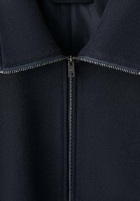 Giacca in misto lana blu navy con colletto alto, presenta una zip a tutta lunghezza e una texture del tessuto liscia. Il dettaglio riflettente della zip aggiunge contrasto.