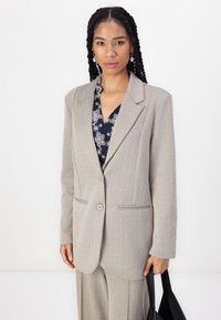 Helehall herringbone bleiser, mille on notšitud kraed, kaks esist pocketit ja üksainus nööp, on koos sinise mustriga pluusiga.